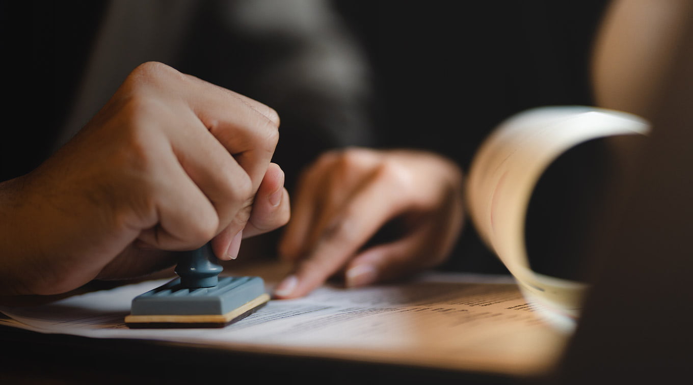 A hand stamps an iGaming compliance document on a dark surface, with a blurred rolled paper in the background.